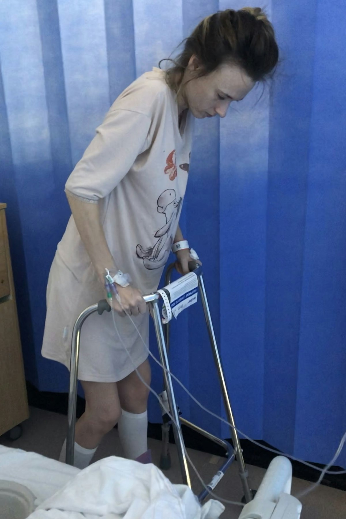 Monica Thomas using a walking frame in a hospital room, wearing a medical gown with IV tubes in her arms, standing beside a blue privacy curtain